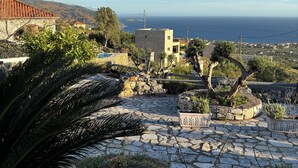 A stone-paved patio with potted plants, a wooden bench, and a view of the sea and distant mountains.