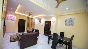 A living room with a sofa, coffee table, and dining table with chairs. There is a ceiling fan, recessed lighting, and a door with a decorative panel. - Executive Two Bedroom Furnished Apartment (Accra)