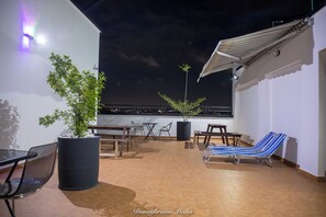 Una terraza en la azotea con una planta en maceta, mobiliario exterior y vistas de la ciudad de noche.