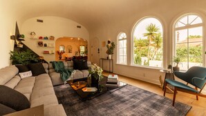 A spacious living room with a large sofa, a coffee table, and a view of a garden through arched windows. - Hauser House, Modern Artful Spanish-style Bungalow (Los Angeles)