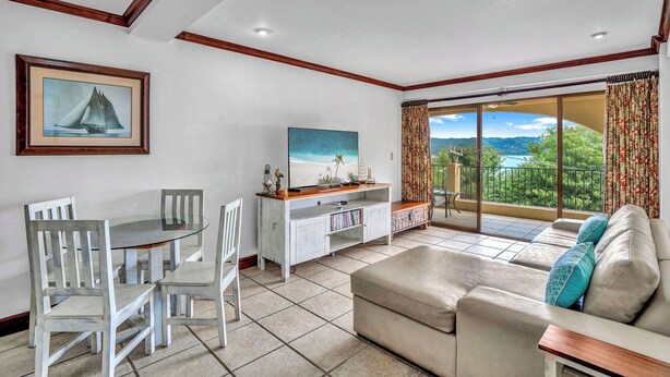 A living room with a dining area, a flat-screen TV, a sofa with cushions, and a balcony with a view of the landscape. - Newly Remodeled Flamingo Condo with Sweeping Ocean Views, Spacious Terrace & Walkable Location (Cabo Velas)
