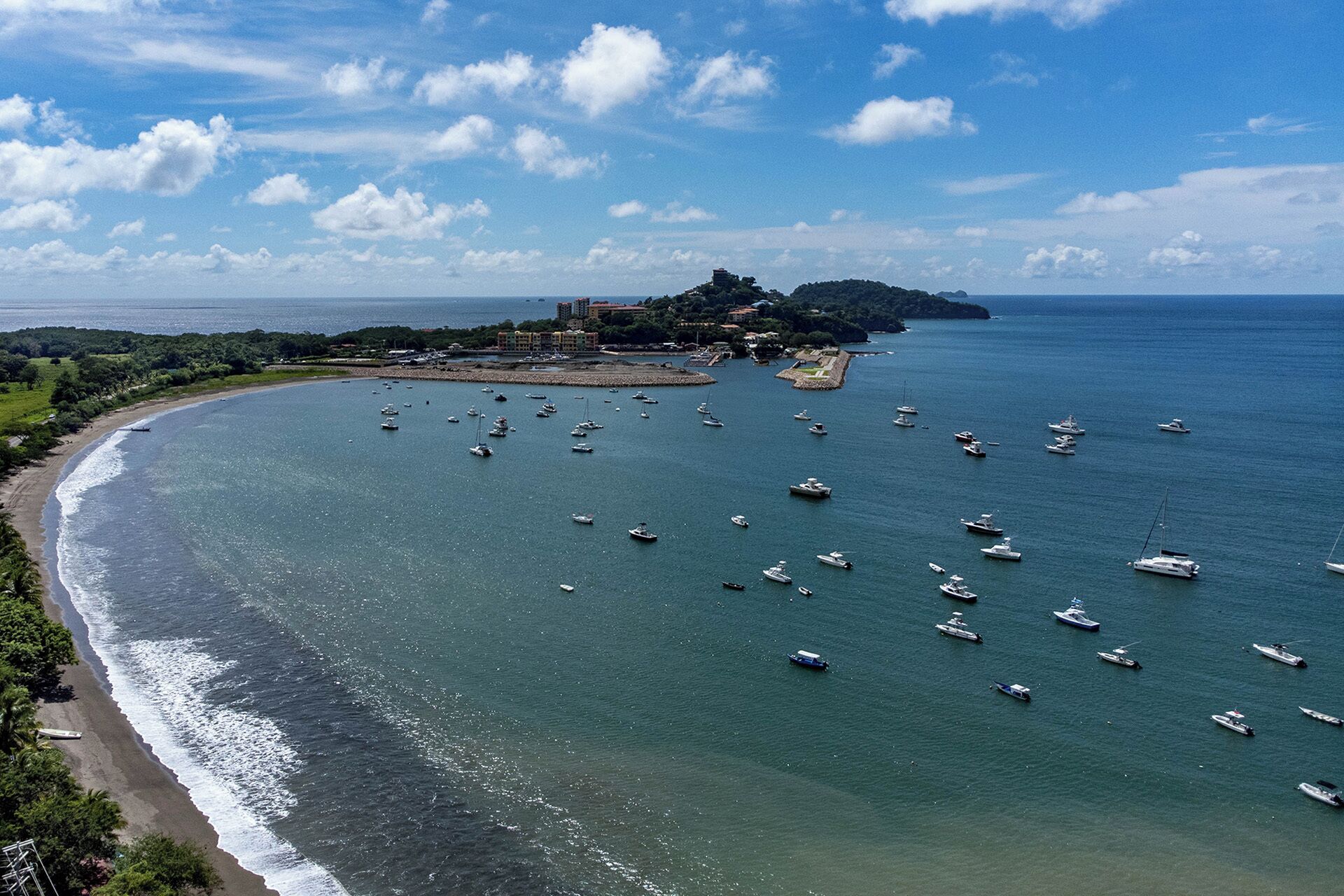 Zona costera con numerosos barcos anclados, una playa y un edificio distante en una colina.