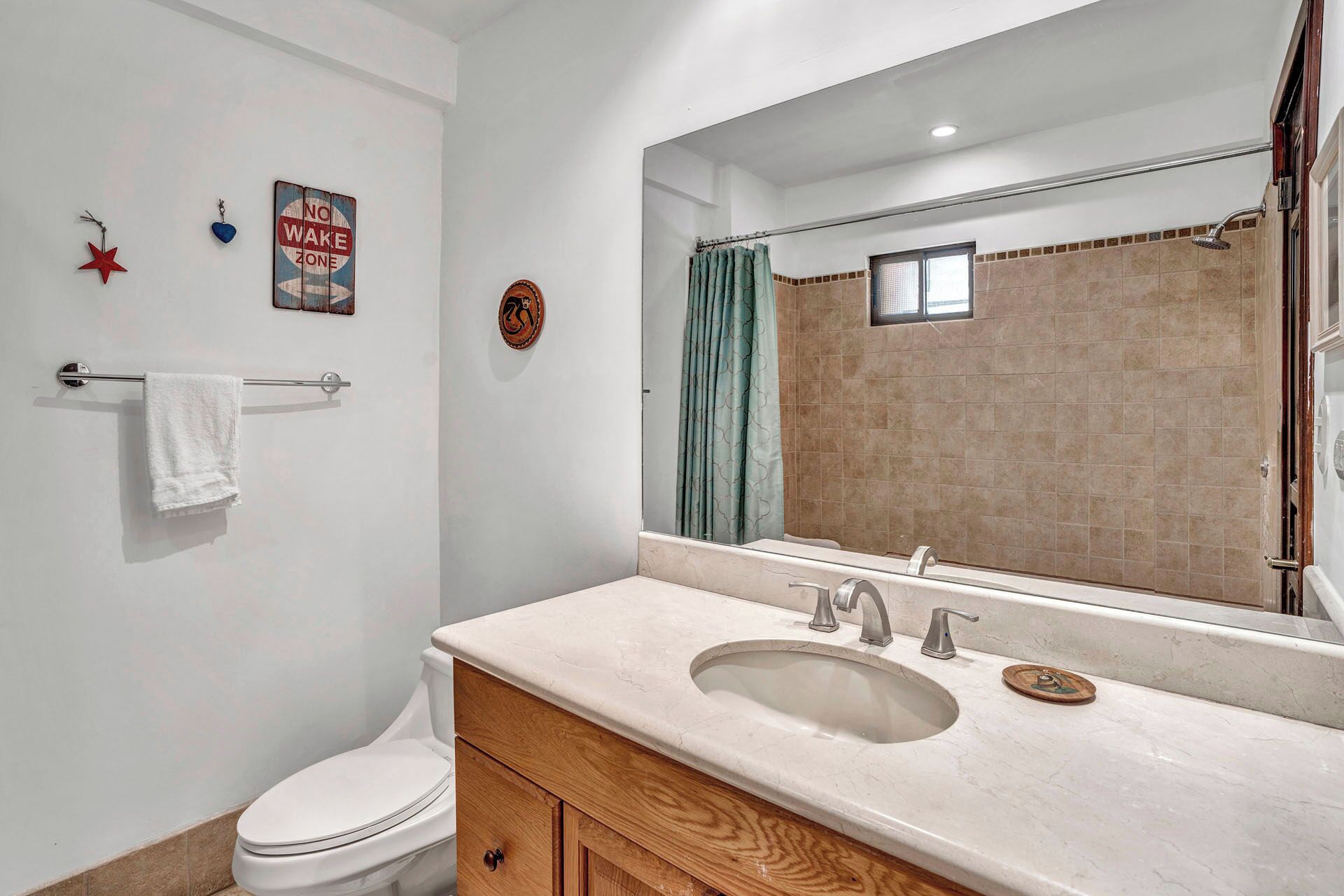 A bathroom with a white sink, vanity, and toilet. There is a towel rack with a white towel, a shower curtain, and a "No Waste Zone" sign on the wall.