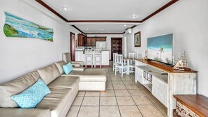 A living room with a sofa, a dining table with chairs, a kitchen area, and a TV. - Newly Remodeled Flamingo Condo with Sweeping Ocean Views, Spacious Terrace & Walkable Location (Cabo Velas)