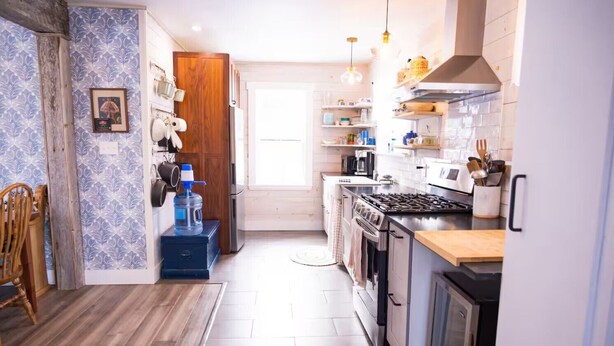 A modern kitchen with a stove, microwave, and various appliances. There is a wooden door and a window with curtains. - Sea la Vie - Margaree farmhouse by river and sea! (Margaree Harbour)