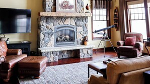 Una sala de estar acogedora con chimenea de piedra, vigas de madera, televisor de pantalla plana, mobiliario de cuero y ventanas amplias.