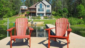 Una propiedad frente al lago con dos sillas Adirondack rojas en una terraza de madera, un muelle que se adentra en un lago tranquilo y una casa grande con balcón con vistas al agua.
