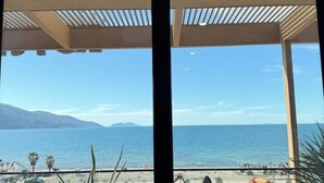Vista de uma varanda coberta, com vista para uma praia, céu claro e montanhas ao longe.