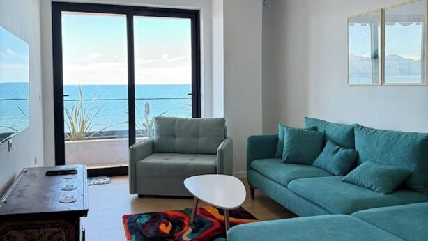 Uma sala de estar moderna com um sofá cinza, um sofá verde-água, uma mesa de centro branca e uma pequena mesa lateral. Há uma grande porta de vidro deslizante que dá para uma varanda com vista para o mar.