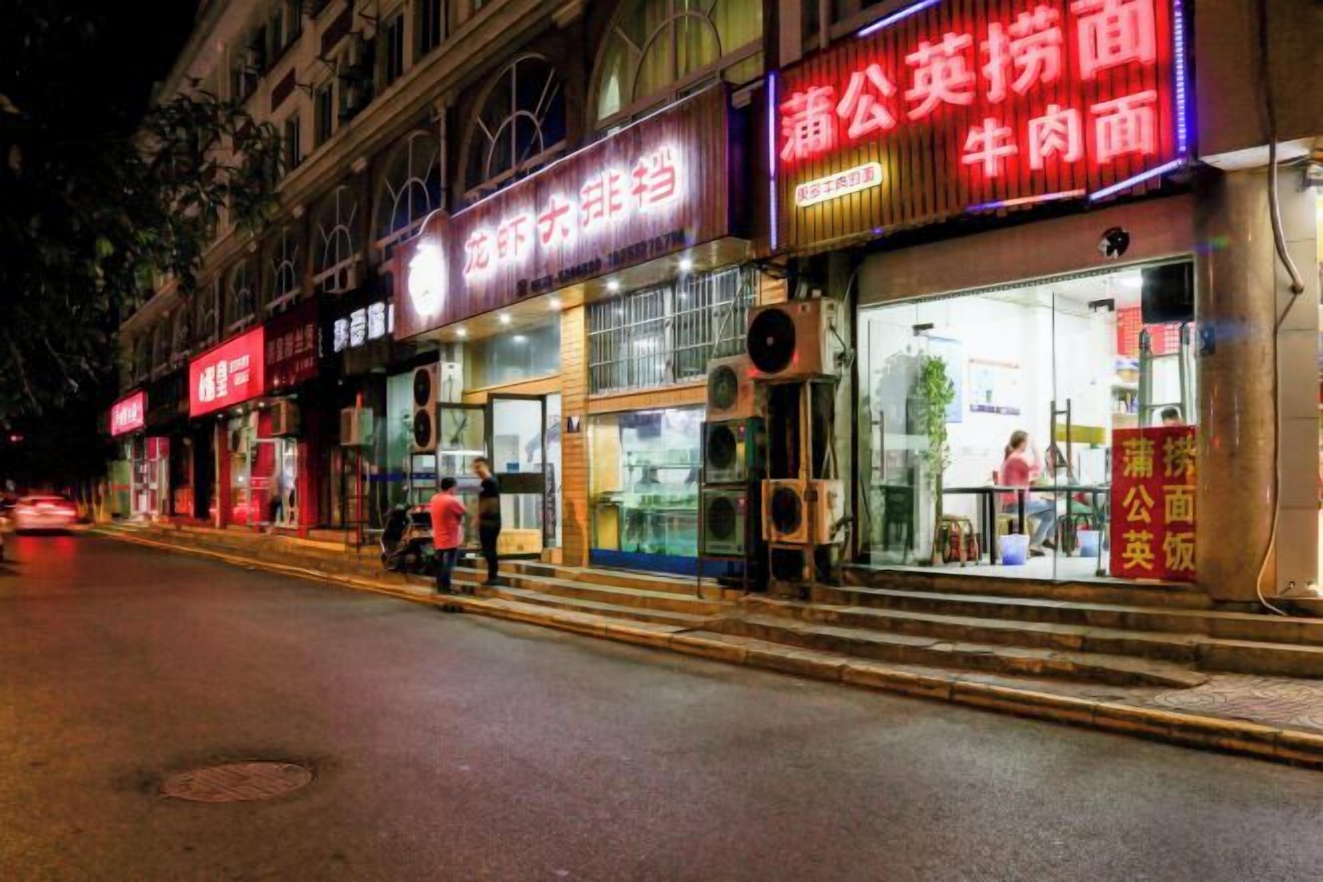 A street view at night with illuminated storefronts, including a restaurant with a glass facade and signage in a foreign language.
