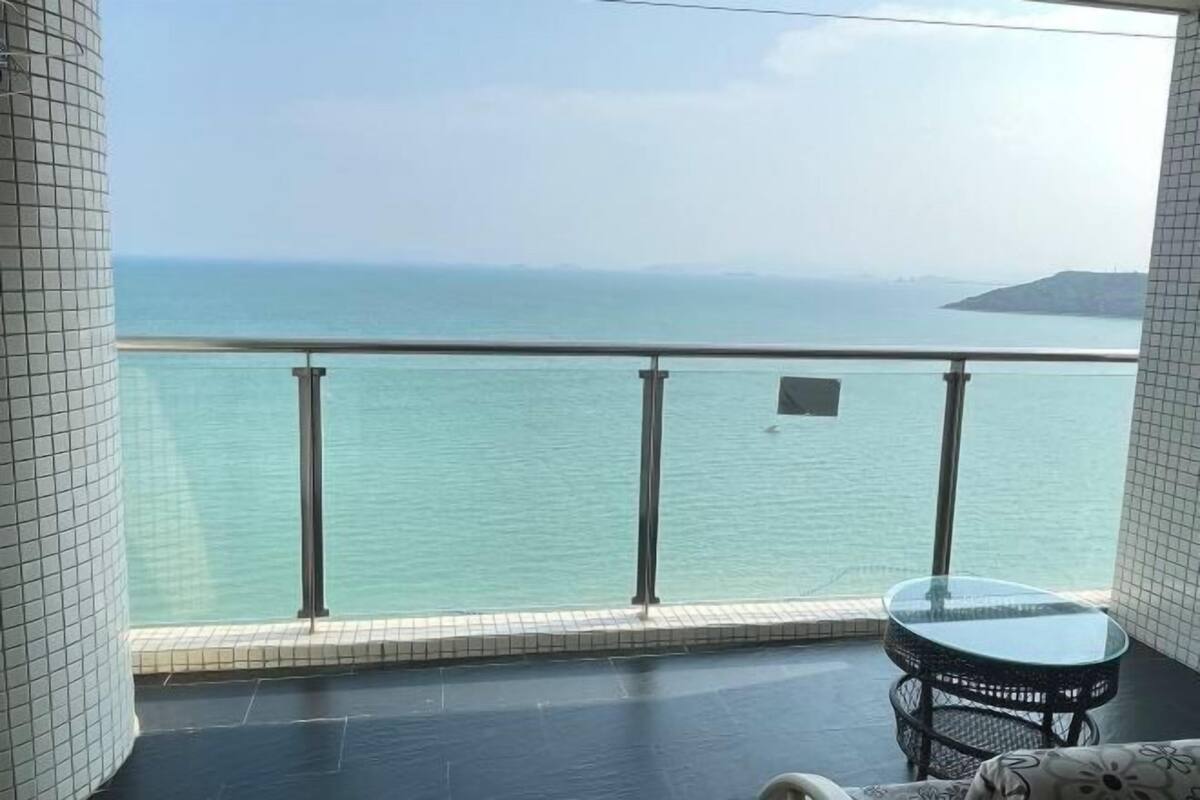 A balcony with a glass table and a chair, overlooking a body of water and distant land.