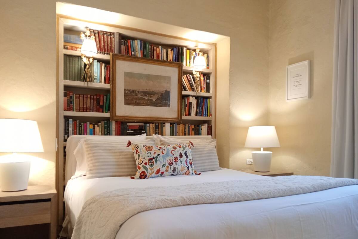 Ein ordentlich eingerichtetes Schlafzimmer mit einem großen Bett, zwei Nachttischen mit Lampen, einer Bücherwand voller Bücher und einem gerahmten Bild an der Wand.