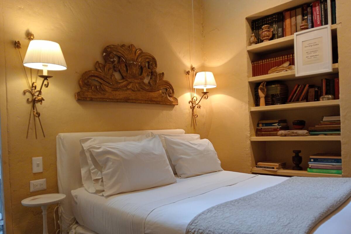 Ein ordentlich bezogenes Bett mit weißen Leinentüchern, zwei Wandleuchten und ein Bücherregal voller Bücher.