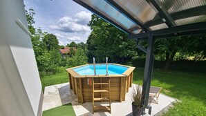 A wooden hot tub with a ladder, surrounded by a patio with artificial grass and a pool.