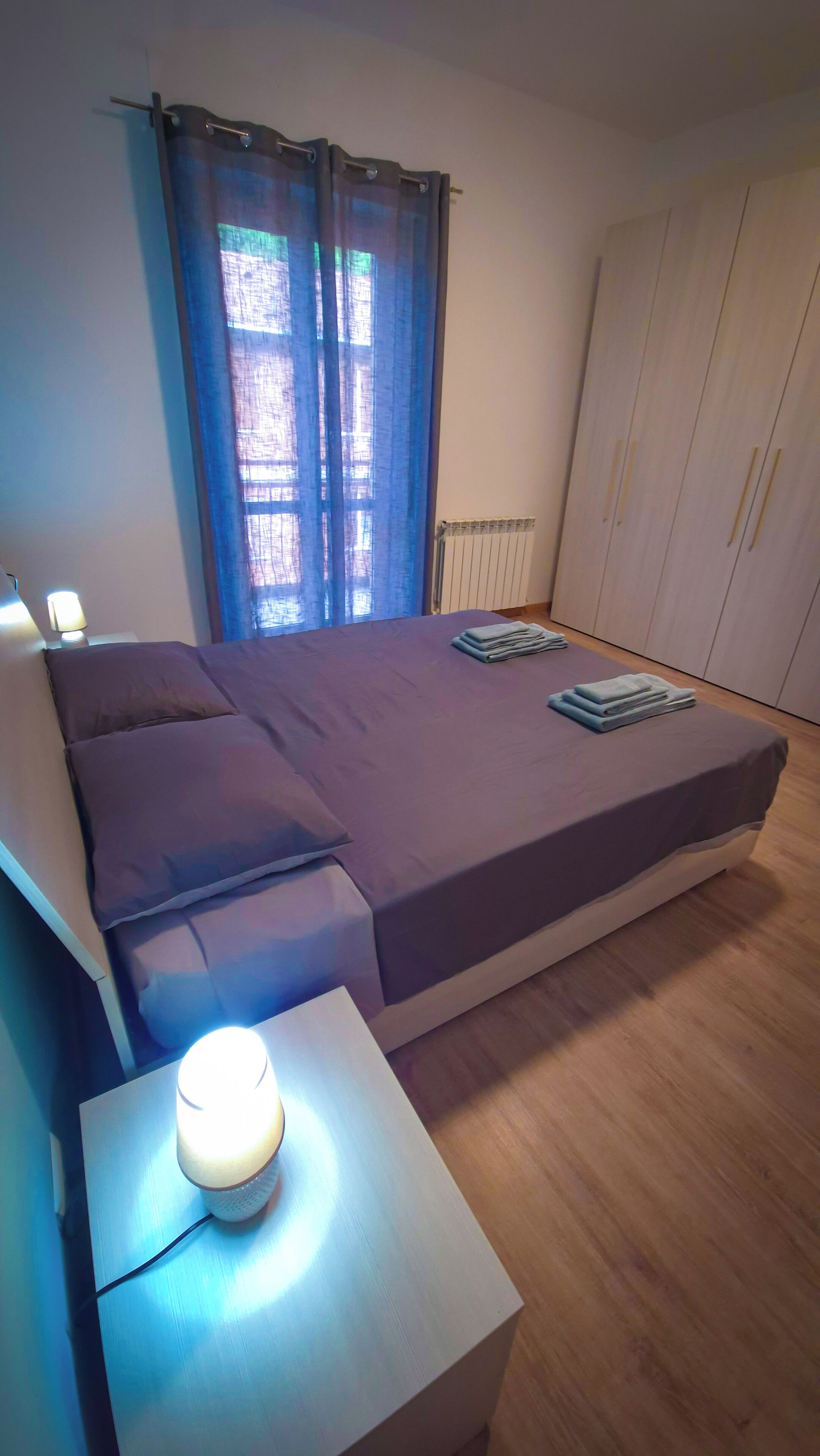 Ein Schlafzimmer mit einem Bett, einem Nachttisch mit Lampe, einem Kleiderschrank und einem Fenster mit Vorhängen.