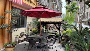 Un área de estar al aire libre con mesa, sillas, un paraguas rojo y plantas en macetas.