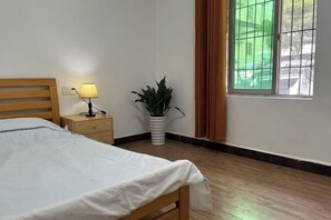 Una cama bien hecha con una colcha blanca, una lámpara de noche, una planta en maceta y una ventana con cortinas.