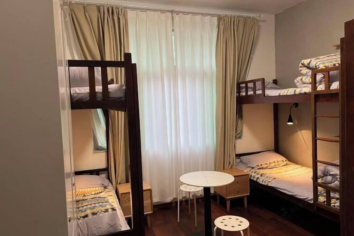 A dormitory room with bunk beds, a small table, and a window with curtains.
