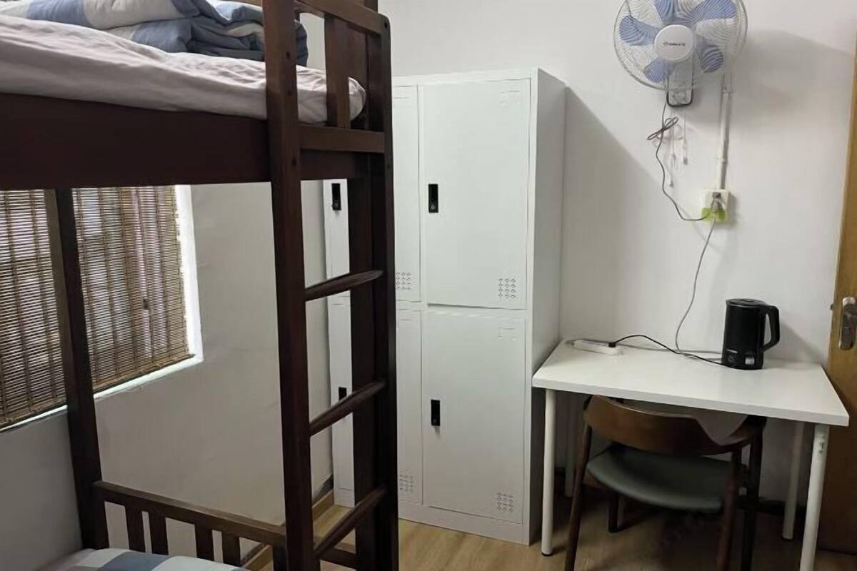 A bunk bed with a window, a desk with a chair, a white wardrobe, and a standing fan.