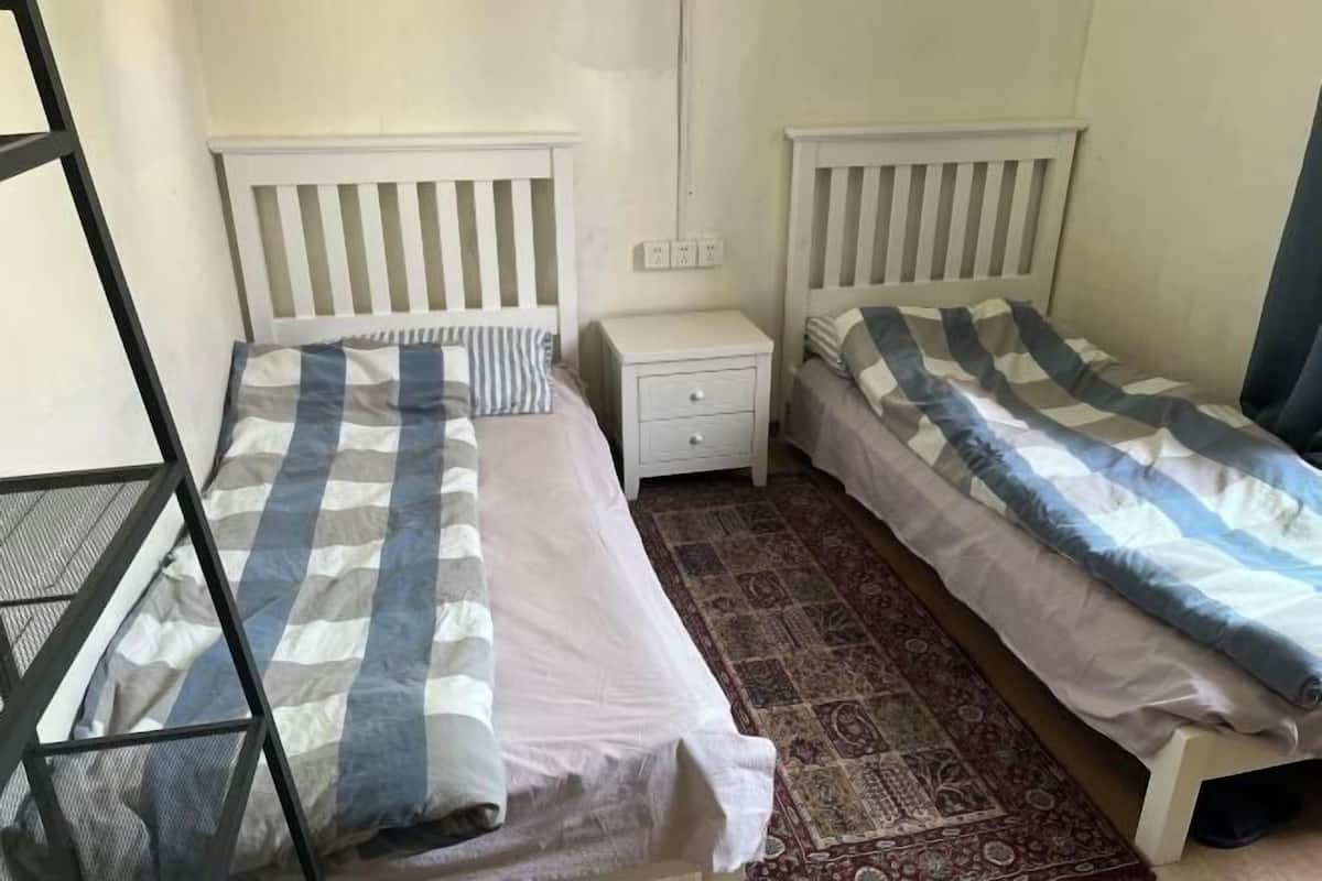 A room with two beds, each with a white headboard, a small bedside table, and a patterned rug on the floor.