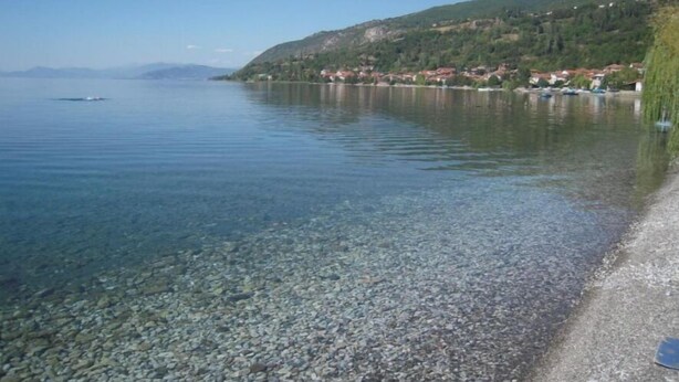 A clear lake with a pebble beach and a hillside with houses.