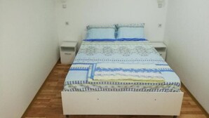 A small, single-bed room with a wooden floor, white walls, and a patterned bedspread. There are two bedside tables and a small white nightstand.