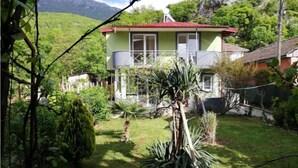 A two-story house with a red roof, surrounded by lush greenery and a mountainous backdrop.