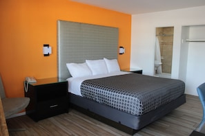 A hotel room with an orange wall, a bed with a grey headboard, a nightstand, a chair, and a shower area. - Torch Lite Lodge  (Yuma)