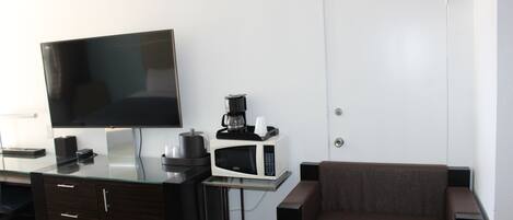 A hotel room with a flat-screen TV, a microwave, a coffee maker, a sofa, a desk with drawers, and a door.
