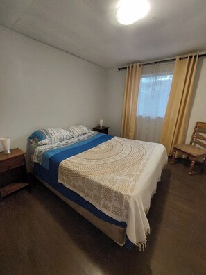 A bedroom with a bed, a nightstand, a chair, and a window with curtains. - Hostal Nahuel Ray (Pucon)