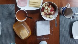 A dining table with a loaf of bread, a plate of ham and cheese, a bowl of fruit, a cup of tea, and a honey jar. - Hostal Nahuel Ray (Pucon)