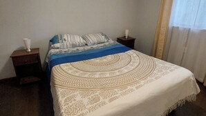 A bedroom with a bed, two bedside tables, a window with curtains, and a plain wall. - Hostal Nahuel Ray (Pucon)