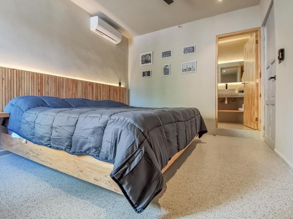 Un dormitorio moderno con cama, cabecera de madera, armario empotrado y un baño pequeño a la vista a través de una puerta abierta.