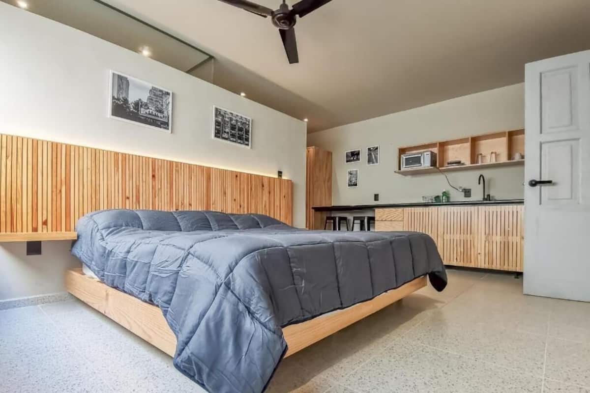 A modern bedroom with a large bed, a wooden headboard, and a kitchen area with a microwave and cabinets.