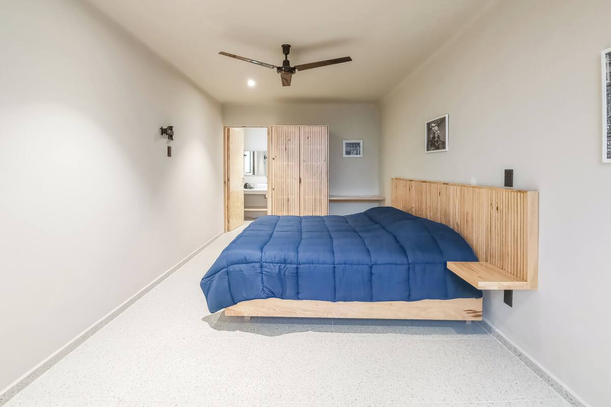 A bedroom with a bed, wooden headboard, ceiling fan, and a view into the bathroom.