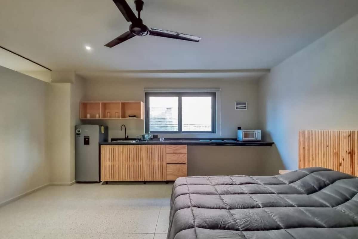 A compact bedroom with a bed, a ceiling fan, a kitchenette with cabinets, and a window.
