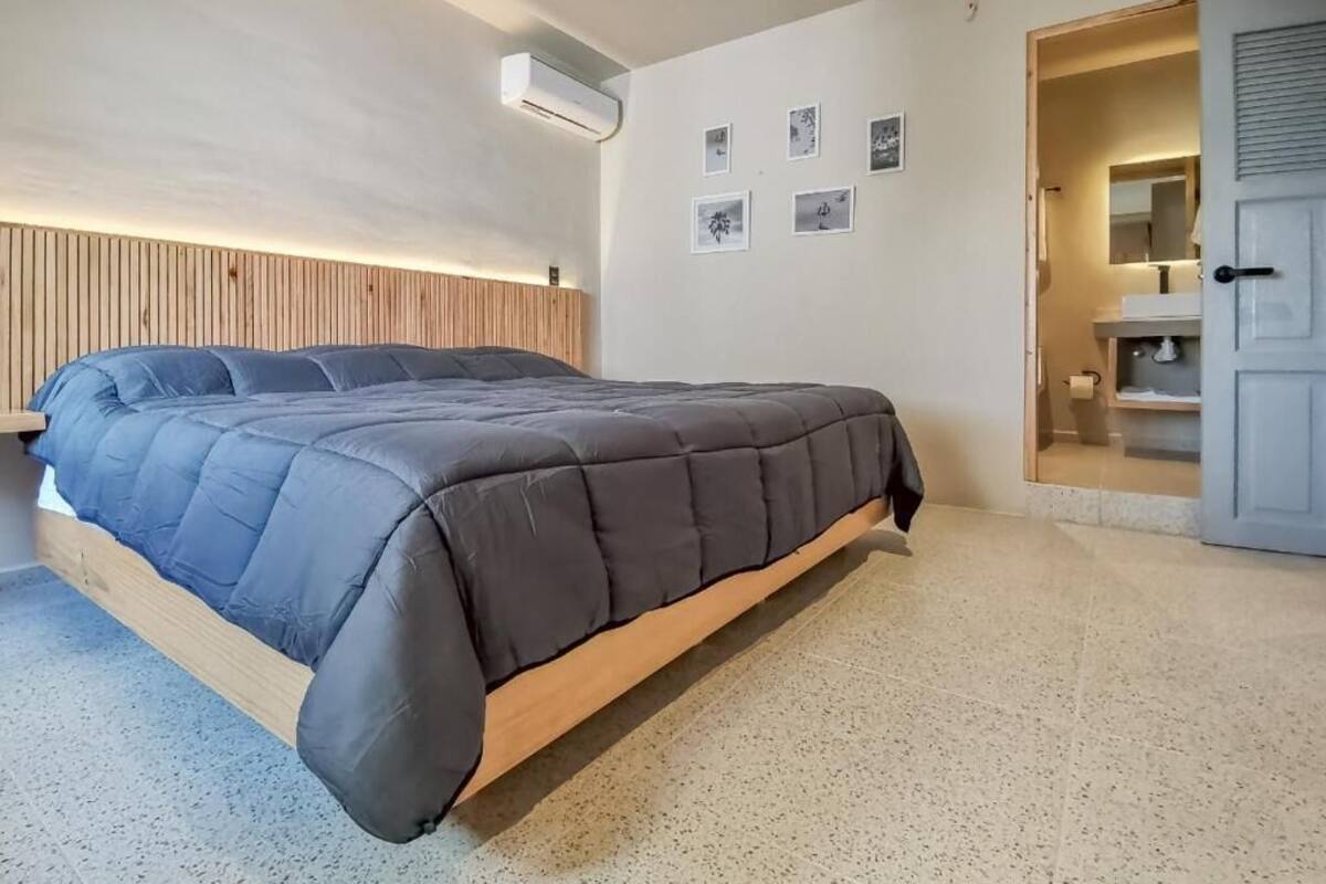 A bedroom with a bed, a wooden headboard, a wall-mounted air conditioner, and a door leading to a bathroom.