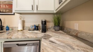 Cozinha com armários brancos, cafeteira, máquina de lavar louça e uma planta em vaso.