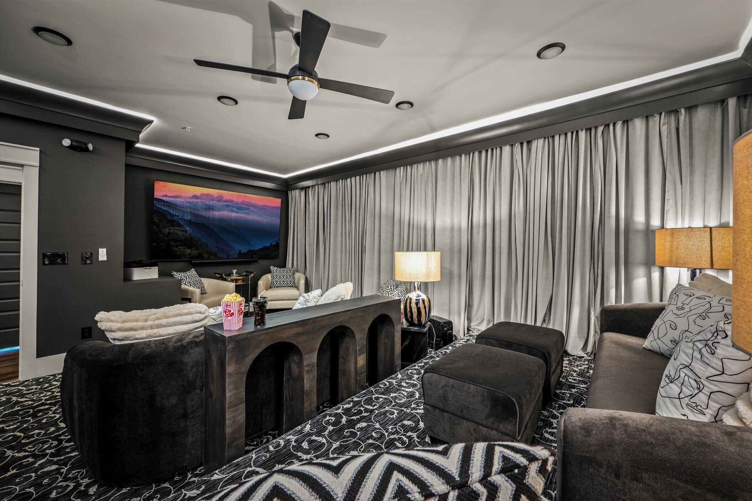 Een moderne woonkamer met een flatscreen televisie, een plafondventilator en een groot raam met gordijnen.