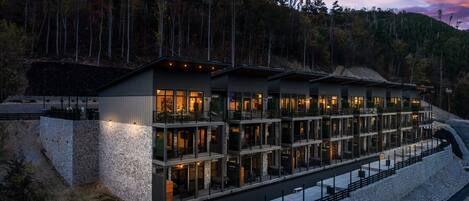 Un edificio moderno de varios pisos con ventanas amplias y balcones, rodeado por un muro de piedra y una ladera boscosa.