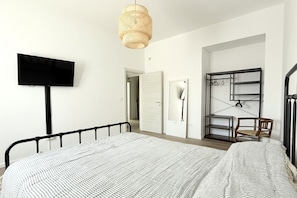 A bedroom with a bed, a television, a hanging light fixture, a wardrobe, and a desk with a chair.