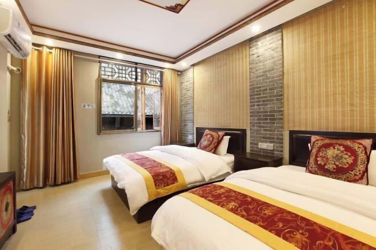 A hotel room with two beds, a large window with curtains, a brick accent wall, and a ceiling fan.