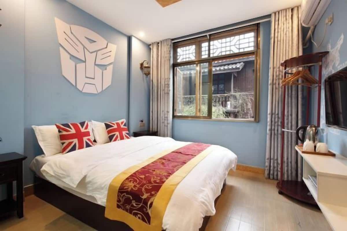 A bedroom with a large bed, a window with curtains, a wall-mounted light, and a small table with a kettle.