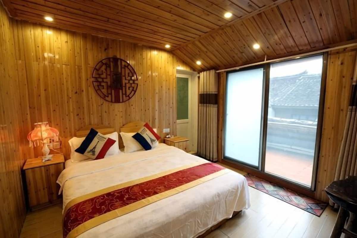 A wooden-paneled bedroom with a large bed, a nightstand, a lamp, and a view of a traditional building through a sliding door.