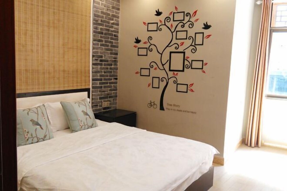 Quarto com cama, decalque de árvore na parede, parede de tijolos e janela com cortinas.