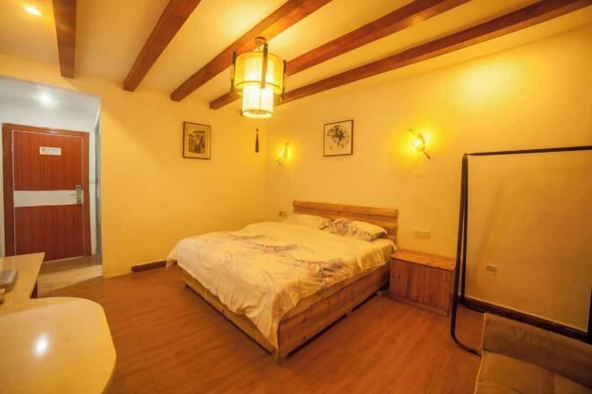 A bedroom with a wooden bed, a nightstand, a desk, and a chair. There are two wall-mounted light fixtures and a door with a sign.