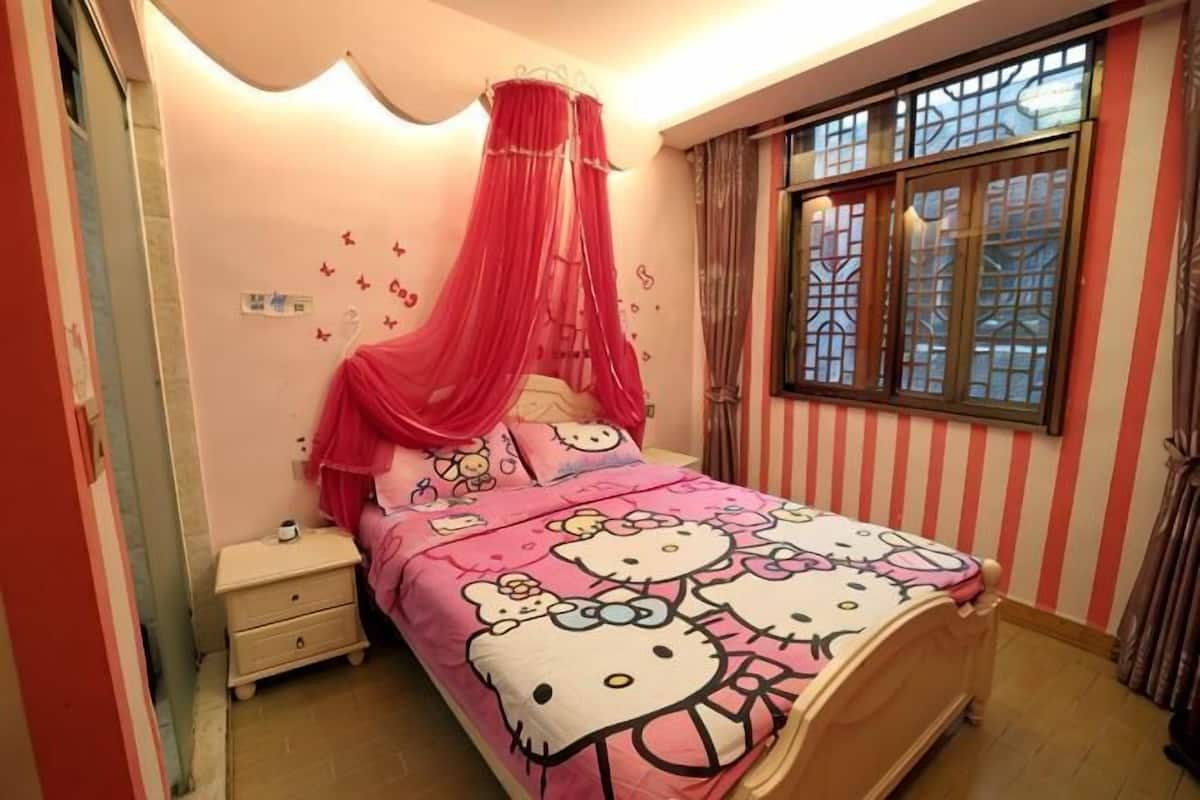 A bed with Hello Kitty bedding, a bedside table, a window with curtains, and a wall with decorative stickers.