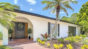 Una casa moderna con un sendero de ladrillo que conduce a una puerta de madera oscura, rodeada de abundante vegetación y una palmera.