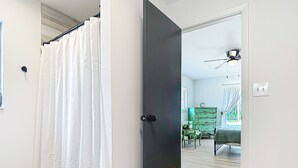 Un pasillo con una puerta oscura, cortinas blancas, piso de madera y vista a una habitación con cama, cómoda y ventilador de techo.