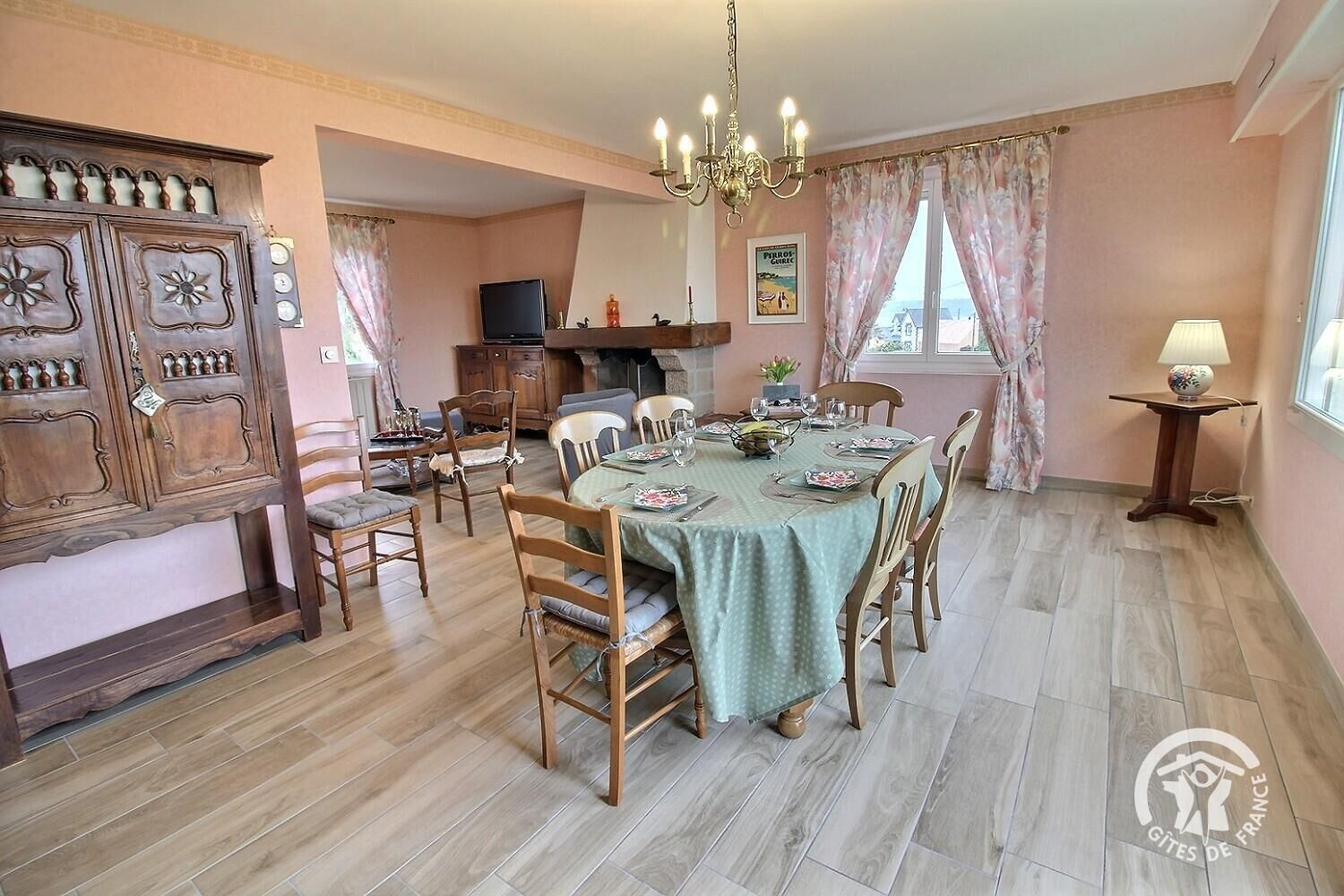A dining area with a table set for four, surrounded by wooden chairs. A chandelier hangs from the ceiling, and a television is mounted on the wall.